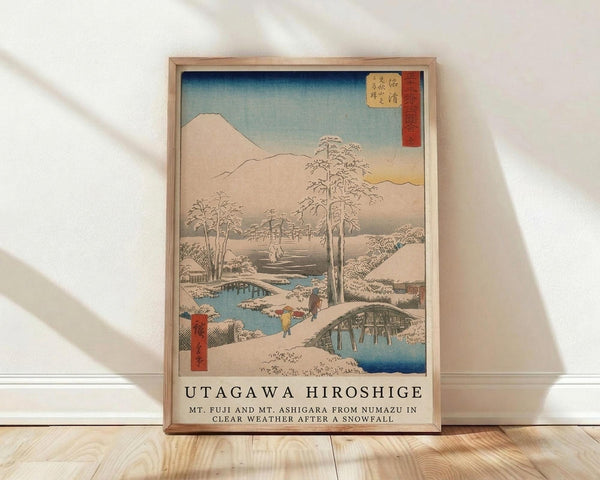 May include: Framed Japanese woodblock print reproduction showing snow-covered winter landscape with Mount Fuji, wooden bridges over blue streams, bare trees, and small figures walking through snowy village. Artwork features soft blues, whites, and earth tones with red text cartouche. Natural wood frame leaning against white wall on light wooden floor with soft shadows.