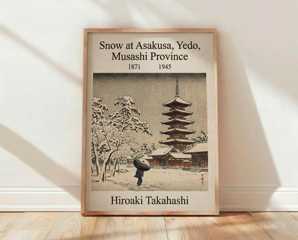 May include: Framed vintage Japanese woodblock print showing a snow-covered temple scene with a five-story pagoda, bare trees laden with snow, and a figure carrying a dark umbrella walking through the white landscape. The artwork features muted earth tones and traditional ukiyo-e style, displayed in a light wooden frame against a white wall on wooden flooring with soft natural lighting creating shadows.