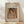 May include: Framed art print displaying a Renoir painting of a woman with dark hair wearing a coral headband and pearl necklace, rendered in soft impressionist brushstrokes with warm peachy tones. The wooden frame sits on light wood flooring against a white textured wall with dramatic palm leaf shadows creating striped patterns across the scene.