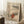 May include: Framed vintage Japanese woodblock print showing figure in red robes walking through snowy mountain landscape with bare tree and pagoda. Print has aged beige background with falling snow. Wooden frame leans against white wall next to white coffee cup on wooden block and cream blanket on textured white rug.