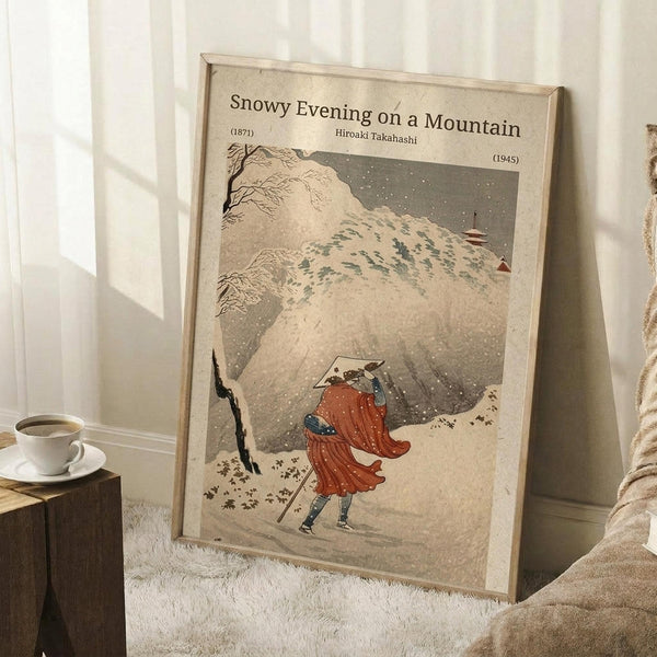May include: Framed vintage Japanese woodblock print showing figure in red robes walking through snowy mountain landscape with bare tree and pagoda. Print has aged beige background with falling snow. Wooden frame leans against white wall next to white coffee cup on wooden block and cream blanket on textured white rug.