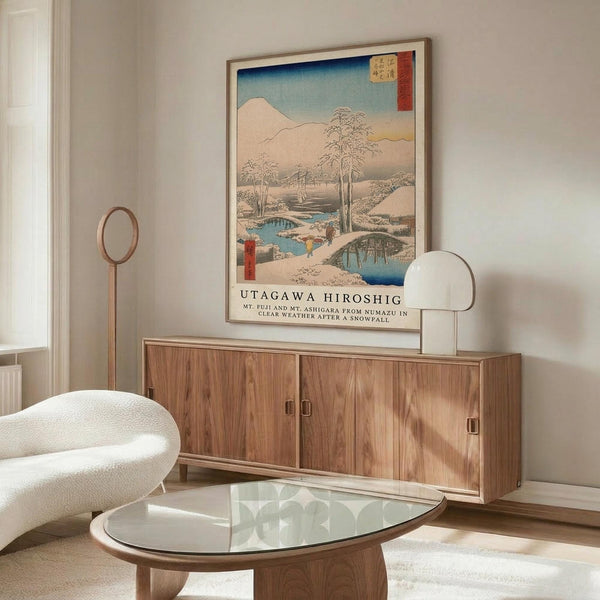 May include: Japanese woodblock print depicting snowy winter landscape with Mount Fuji, bridges over blue water, and bare trees, displayed in thin wooden frame above mid-century modern walnut credenza. White sculptural chair and glass-top coffee table visible in minimalist living room with cream walls and natural lighting.