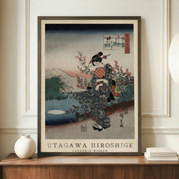 May include: Framed Japanese woodblock print showing a woman in dark patterned kimono with red obi sash, surrounded by cherry blossoms against mountain landscape backdrop. Print displays in modern frame on wooden credenza beside white ceramic vase and stacked books in neutral-toned room with cream walls and white trim molding.