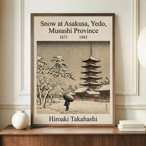 May include: Framed vintage Japanese woodblock print showing snow-covered temple pagoda with figure holding umbrella, displayed on wooden console beside white ceramic vase and books against cream wall with white molding