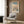 May include: Japanese woodblock print artwork displayed in modern living room with beige armchair, gold floor lamp, and wood paneling. The framed poster shows a winter landscape with snow-covered mountains, bare trees, wooden bridge over blue water, and small figures walking through the snowy scene. Soft natural lighting from window illuminates the neutral-toned space with geometric patterned throw pillow.
