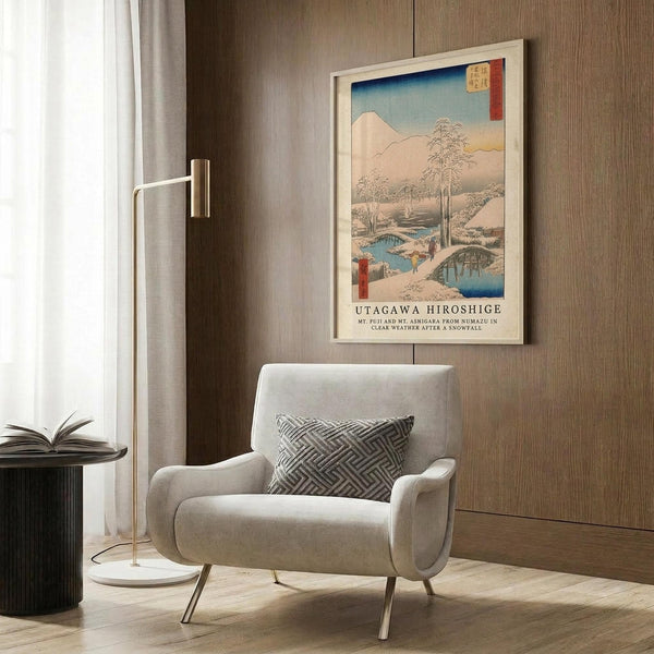 May include: Japanese woodblock print artwork displayed in modern living room with beige armchair, gold floor lamp, and wood paneling. The framed poster shows a winter landscape with snow-covered mountains, bare trees, wooden bridge over blue water, and small figures walking through the snowy scene. Soft natural lighting from window illuminates the neutral-toned space with geometric patterned throw pillow.