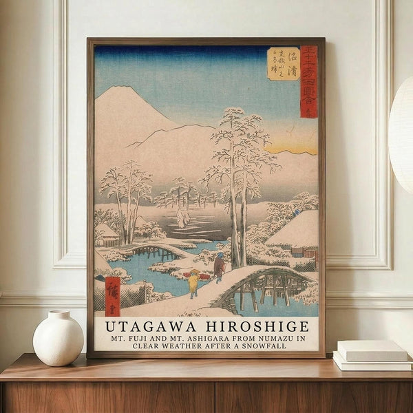 May include: Framed Japanese woodblock print depicting snowy winter landscape with Mount Fuji, snow-covered trees, wooden bridges over blue stream, and small figures walking. Print features soft blue sky gradient, muted earth tones, and traditional red text cartouche. Displayed on wooden sideboard beside white ceramic vase and books in elegant room with white paneled walls.