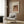 May include: Framed vintage art print featuring a woman with dark hair in soft pink and coral tones, displayed on wood paneled wall above a cream upholstered armchair with geometric patterned pillow. Modern reading corner with brass floor lamp, black side table with open book, and natural lighting from window with white curtains.