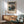May include: Framed vintage Japanese woodblock print showing woman in ornate blue kimono with floral patterns against mountain landscape backdrop, displayed above light wood sideboard with white ceramic lamp, next to sage green mid-century armchair with wooden arms in bright sunlit room with white walls.