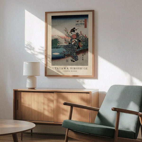 May include: Framed vintage Japanese woodblock print showing woman in ornate blue kimono with floral patterns against mountain landscape backdrop, displayed above light wood sideboard with white ceramic lamp, next to sage green mid-century armchair with wooden arms in bright sunlit room with white walls.