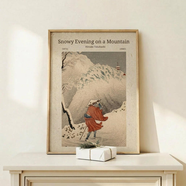 May include: Framed vintage Japanese woodblock print showing a figure in red robes walking through heavy snowfall on a mountain path, with snow-covered trees and buildings visible. The artwork has muted earth tones and is displayed in a light wooden frame on a white wooden surface next to a small wrapped gift with twine and greenery.