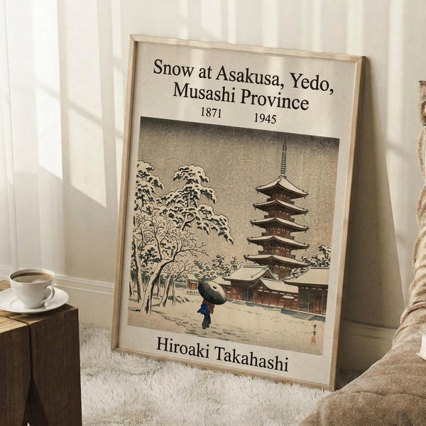 May include: A framed Japanese woodblock print poster leaning against a white wall, showing a snow-covered temple pagoda and figure with umbrella in muted earth tones. The beige wooden frame sits on a white textured rug beside a coffee cup on a wooden block and cream blanket.