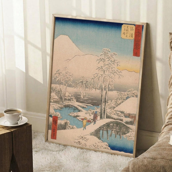 May include: Japanese woodblock print in light wood frame leaning against white wall, depicting snowy winter landscape with Mount Fuji, wooden bridge over blue stream, bare trees, and small figures in colorful clothing. Red cartouche with Japanese text in upper right corner. Coffee cup on wooden block nearby, white textured rug below, cozy indoor setting with natural lighting.