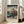 May include: Framed vintage Japanese woodblock print featuring a woman in ornate kimono with floral patterns, set against mountain landscape with cherry blossoms. Light wooden frame displays the artwork on wooden floor next to beige upholstered chair in bright, minimalist room with white walls and large windows.
