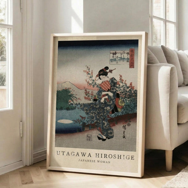 May include: Framed vintage Japanese woodblock print featuring a woman in ornate kimono with floral patterns, set against mountain landscape with cherry blossoms. Light wooden frame displays the artwork on wooden floor next to beige upholstered chair in bright, minimalist room with white walls and large windows.