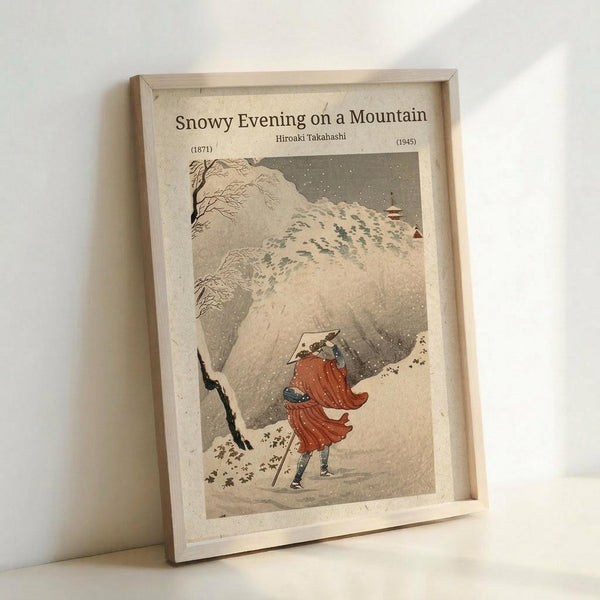 May include: Framed vintage Japanese woodblock print showing a figure in red robes and conical hat walking through heavy snow on a mountain path, carrying a walking stick. Snow-covered trees and buildings visible in background. Print has muted earth tones with falling snow effect. Displayed in natural wood frame leaning against white wall.