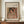 May include: Framed poster print of a Renoir painting featuring a woman with dark hair and flowing fabric in warm peachy tones, displayed on a wooden sideboard next to a cream ceramic vase and white books in an elegant room with cream walls and wainscoting.