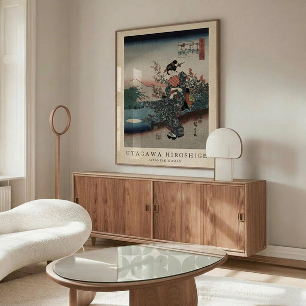 May include: A framed Japanese woodblock print featuring a woman in traditional dress with Mount Fuji in the background, displayed above a mid-century modern wooden credenza with sliding doors. The room includes a white curved chair, glass-top oval coffee table, and modern floor lamp, creating a minimalist aesthetic with warm wood tones against neutral walls.