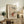 May include: Framed vintage Japanese woodblock print showing figure in red coat walking through snowy mountain landscape with falling snow. Print displayed on rustic wooden sideboard alongside fern plant, books, ceramic vase and table lamp in neutral-toned living room setting with beige upholstered chair.