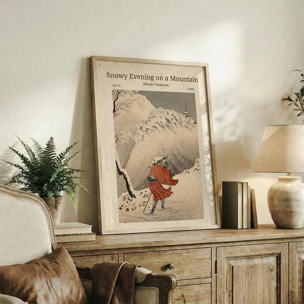 May include: Framed vintage Japanese woodblock print showing figure in red coat walking through snowy mountain landscape with falling snow. Print displayed on rustic wooden sideboard alongside fern plant, books, ceramic vase and table lamp in neutral-toned living room setting with beige upholstered chair.