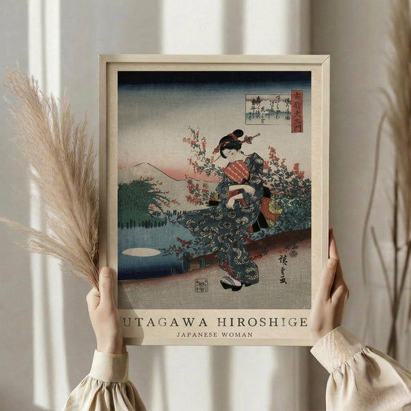 May include: Hands holding a framed reproduction of a Japanese woodblock print showing a woman in traditional kimono with floral patterns standing among cherry blossoms and mountains. The artwork features muted colors with blue sky gradient, pink flowers, and traditional Japanese artistic style. Natural light wooden frame against neutral background with dried pampas grass visible on left side.