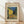 May include: Framed poster print of Van Gogh's famous cafe terrace night scene showing yellow building with outdoor seating, cobblestone street, and starry blue sky. Light wood frame with cream matting, leaning against white wall on wooden floor with soft natural lighting creating shadows.