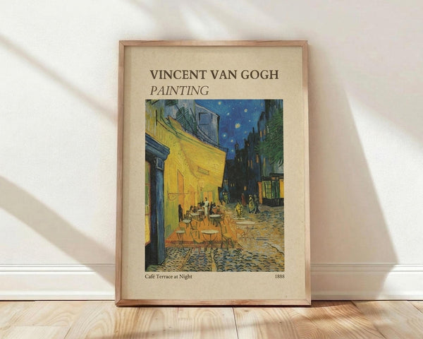 May include: Framed poster print of Van Gogh's famous cafe terrace night scene showing yellow building with outdoor seating, cobblestone street, and starry blue sky. Light wood frame with cream matting, leaning against white wall on wooden floor with soft natural lighting creating shadows.