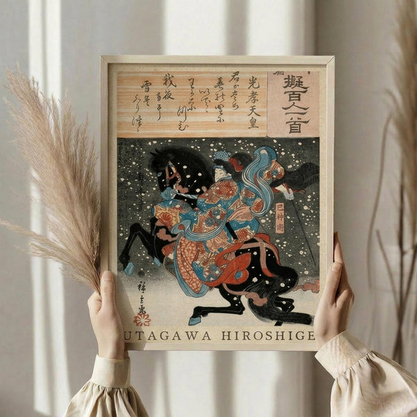 May include: Hands holding a framed Japanese woodblock print showing a warrior in colorful robes battling in falling snow. The artwork features traditional ukiyo-e style with Japanese calligraphy at top, vibrant blues and oranges, and dramatic action scene. Light wood frame against white curtained background with dried pampas grass visible on left side.