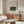 May include: Framed art print of Henri Rousseau's jungle painting featuring a lion attacking an antelope in lush green foliage, displayed above a walnut mid-century modern credenza with brass handles. Modern living room setting with cream upholstered swivel chair, glass-top oval coffee table, white marble lamp, and brass floor mirror. Neutral walls and natural lighting create minimalist Scandinavian aesthetic.