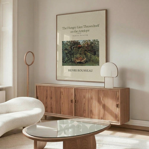 May include: Framed art print of Henri Rousseau's jungle painting featuring a lion attacking an antelope in lush green foliage, displayed above a walnut mid-century modern credenza with brass handles. Modern living room setting with cream upholstered swivel chair, glass-top oval coffee table, white marble lamp, and brass floor mirror. Neutral walls and natural lighting create minimalist Scandinavian aesthetic.