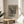 May include: Black and white reproduction of Edvard Munch's The Scream artwork displayed in thin wood frame on round wooden side table next to beige upholstered chair. Poster shows iconic figure with hands to face on bridge with swirling sky, includes text identifying it as masterpiece of expressionism. Soft natural lighting creates shadows across neutral-toned modern interior with plant visible in corner.
