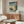 May include: Japanese woodblock print poster featuring ships with colorful sails on blue-green waters near a coastal cliff with pine trees, displayed in thin gold frame above modern walnut sideboard. White curved chair and glass oval coffee table visible in foreground of bright, minimalist room with white walls and polished floors.