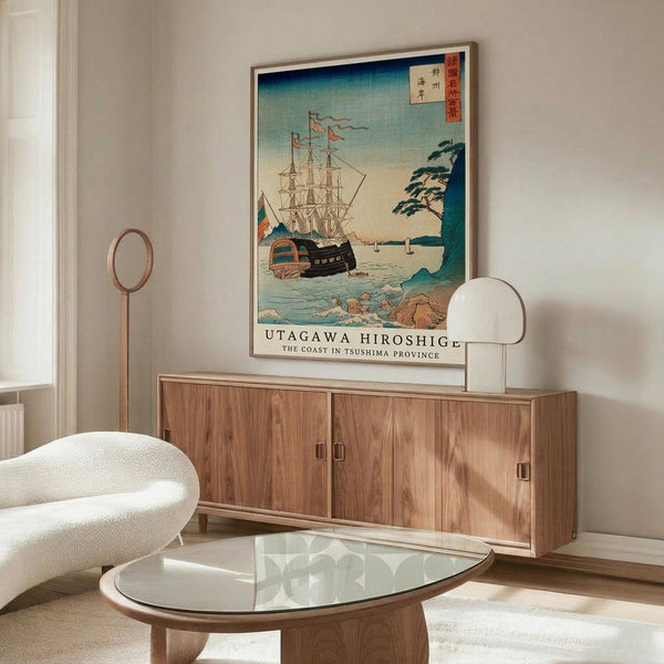 May include: Japanese woodblock print poster featuring ships with colorful sails on blue-green waters near a coastal cliff with pine trees, displayed in thin gold frame above modern walnut sideboard. White curved chair and glass oval coffee table visible in foreground of bright, minimalist room with white walls and polished floors.