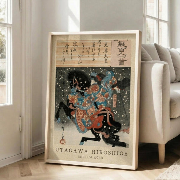 May include: Framed Japanese woodblock print artwork leaning against white wall next to beige sofa in bright room with large windows. The vintage-style poster features a figure in ornate blue and orange robes amid swirling snow or petals against dark background, with Japanese text above and English text below. Light wood frame, approximately 2-3 feet tall, positioned on wooden floor beside furniture leg.