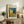 May include: Framed Van Gogh reproduction poster of Cafe Terrace at Night displayed on rustic wooden sideboard alongside fern plant, books, ceramic vase and table lamp. The artwork shows the famous yellow cafe terrace under starry blue night sky with cobblestone street. Light wood frame complements the warm, neutral-toned living room setting with cream upholstered chair and brown throw pillows.