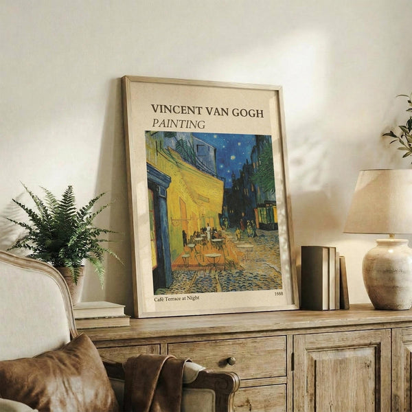May include: Framed Van Gogh reproduction poster of Cafe Terrace at Night displayed on rustic wooden sideboard alongside fern plant, books, ceramic vase and table lamp. The artwork shows the famous yellow cafe terrace under starry blue night sky with cobblestone street. Light wood frame complements the warm, neutral-toned living room setting with cream upholstered chair and brown throw pillows.