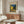 May include: Van Gogh's Cafe Terrace at Night poster print displayed above a mid-century modern walnut credenza in a minimalist living room. The vintage-style poster features the iconic yellow cafe scene with blue starry sky, framed in thin brown frame against white wall. Room includes white curved chair, glass oval coffee table, and warm natural lighting creating soft shadows on light wood furniture.