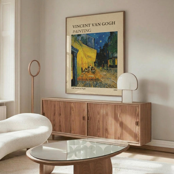 May include: Van Gogh's Cafe Terrace at Night poster print displayed above a mid-century modern walnut credenza in a minimalist living room. The vintage-style poster features the iconic yellow cafe scene with blue starry sky, framed in thin brown frame against white wall. Room includes white curved chair, glass oval coffee table, and warm natural lighting creating soft shadows on light wood furniture.