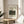 May include: Framed art print displaying Henri Rousseau's jungle painting with a lion attacking an antelope among dense green foliage. The print sits on a light wooden round side table next to a beige upholstered chair. Natural sunlight streams across the neutral-toned room, casting soft shadows on white walls. A plant's leaves are visible in the foreground, creating an organic, minimalist home decor setting.