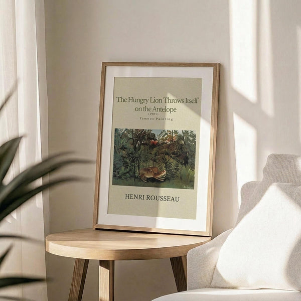 May include: Framed art print displaying Henri Rousseau's jungle painting with a lion attacking an antelope among dense green foliage. The print sits on a light wooden round side table next to a beige upholstered chair. Natural sunlight streams across the neutral-toned room, casting soft shadows on white walls. A plant's leaves are visible in the foreground, creating an organic, minimalist home decor setting.