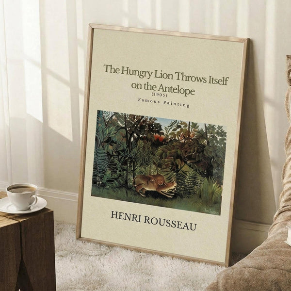 May include: Framed art print leaning against white wall on white textured rug. Print shows Henri Rousseau's jungle painting with lion attacking antelope amid lush green foliage. Cream-colored poster with black text and thin wooden frame. Coffee cup on wooden block visible to left, beige blanket draped nearby. Soft natural lighting creates gentle shadows on wall.