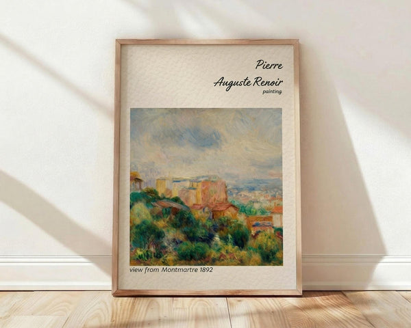 May include: Framed art print of Pierre Auguste Renoir's 1892 Impressionist landscape painting showing view from Montmartre with colorful buildings, lush green foliage, and cloudy sky in warm earth tones. Natural wood frame sits on light wooden floor against white wall with soft window shadows.