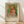 May include: Framed art print featuring a young girl with blonde curly hair wearing a white dress with red vest, painted in soft impressionist style with green and yellow background. The poster displays "PIERRE AUGUSTE exhibition" text below the artwork with "1904" and "Claude Renoir" attribution. Wooden frame rests against white wall on light wood floor with natural sunlight casting diagonal shadows across the scene.