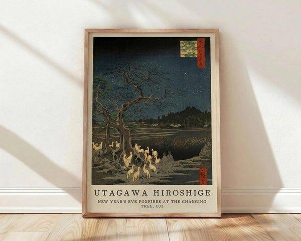 May include: Framed Japanese woodblock print reproduction showing foxfires gathering under a bare tree at night. The artwork features a dark blue starry sky, ghostly white fox spirits beneath twisted branches, and distant hills with traditional buildings. The print has characteristic ukiyo-e style with muted earth tones and includes red Japanese text cartouches. Artist and title text appears below the image on a cream background. The wooden frame leans against a white wall on light wood flooring