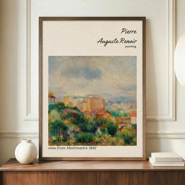 May include: Framed reproduction of Renoir's impressionist landscape painting showing rolling hills with lush green vegetation, colorful buildings, and soft cloudy sky in warm earth tones. Print displayed on wooden sideboard beside white ceramic vase and stacked books against cream paneled wall. Thin brown frame with white matting surrounds the artwork.
