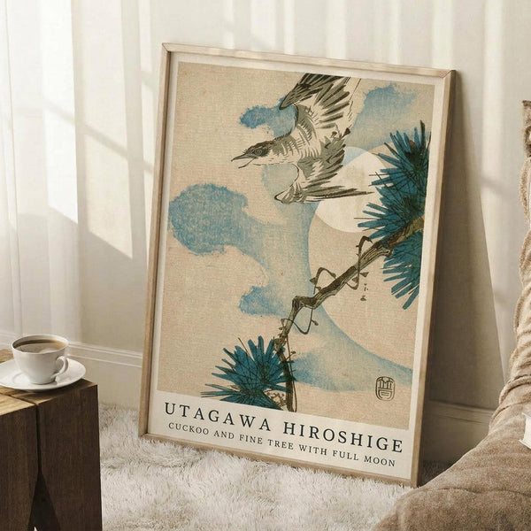 May include: Framed Japanese woodblock print showing a cuckoo bird in flight among pine branches and blue clouds against a cream background. The artwork is displayed in a light wooden frame leaning against white walls in a cozy room setting with a coffee cup on a wooden block, white fluffy rug, and soft natural lighting creating geometric shadows.