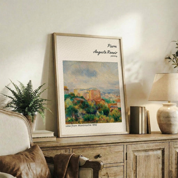May include: Framed Renoir reproduction showing impressionist landscape with lush green foliage, red-orange buildings, and soft cloudy sky in muted pastels. Light wood frame with white matting displays against cream wall on rustic wooden sideboard alongside fern plant, ceramic vase, and stacked books in warm, naturally-lit living space.