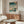 May include: Japanese woodblock print poster in thin wooden frame hanging above walnut credenza in modern living room. Artwork shows winter scene with snow, figures with umbrellas crossing bridge, turquoise sky with falling snow, traditional architecture. White sculptural chair, glass oval coffee table, and minimalist table lamp visible. Clean contemporary interior with white walls and natural lighting.