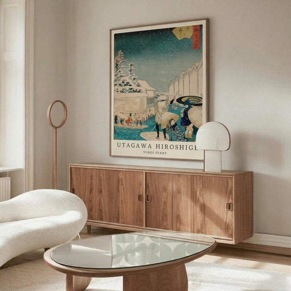 May include: Japanese woodblock print poster in thin wooden frame hanging above walnut credenza in modern living room. Artwork shows winter scene with snow, figures with umbrellas crossing bridge, turquoise sky with falling snow, traditional architecture. White sculptural chair, glass oval coffee table, and minimalist table lamp visible. Clean contemporary interior with white walls and natural lighting.