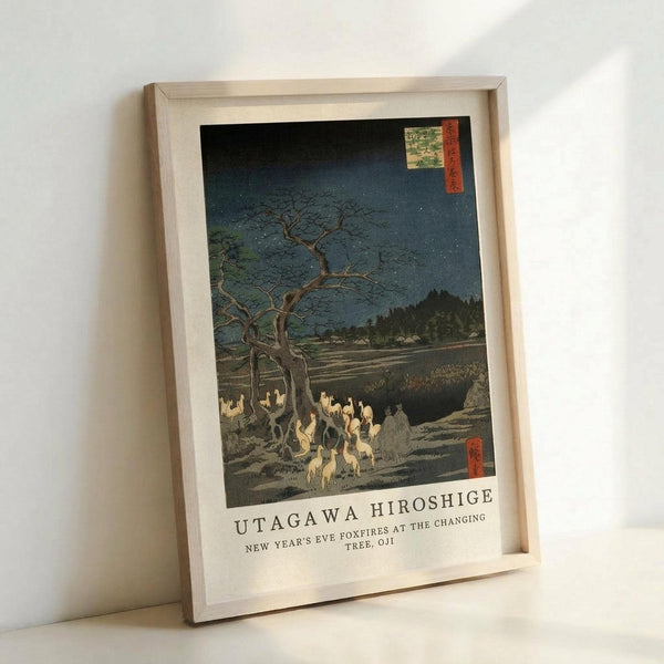 May include: A framed vintage Japanese woodblock print showing foxes with glowing flames gathered under a bare tree at night. The artwork features a dark blue starry sky, barren landscape, and traditional Japanese text cartouches in the corners. The print is displayed in a light wood frame with white matting, leaning against a white wall on a white surface.